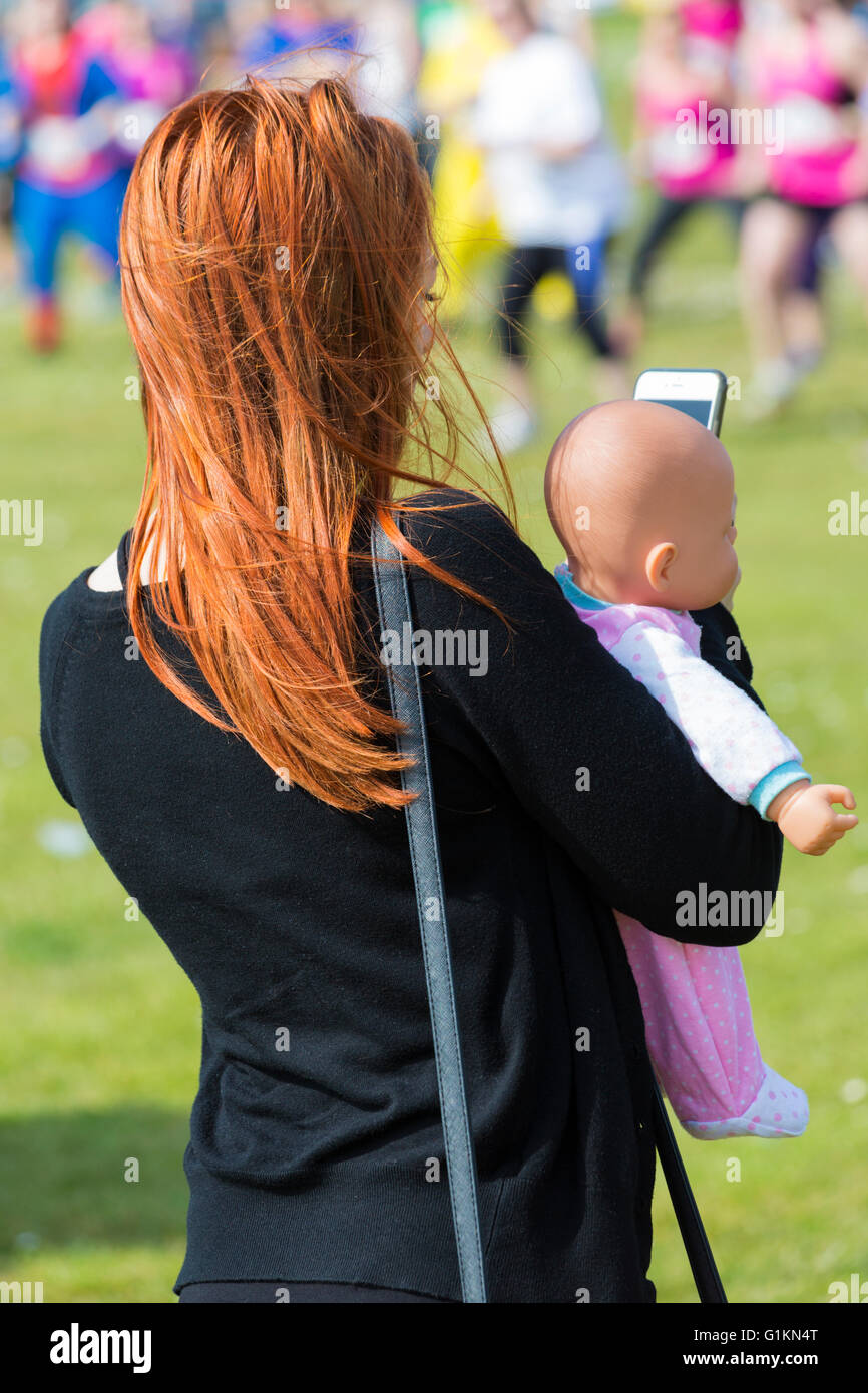 Young woman with long ginger hair holding baby doll whilst looking at phone at event on Poole Stock Photo
