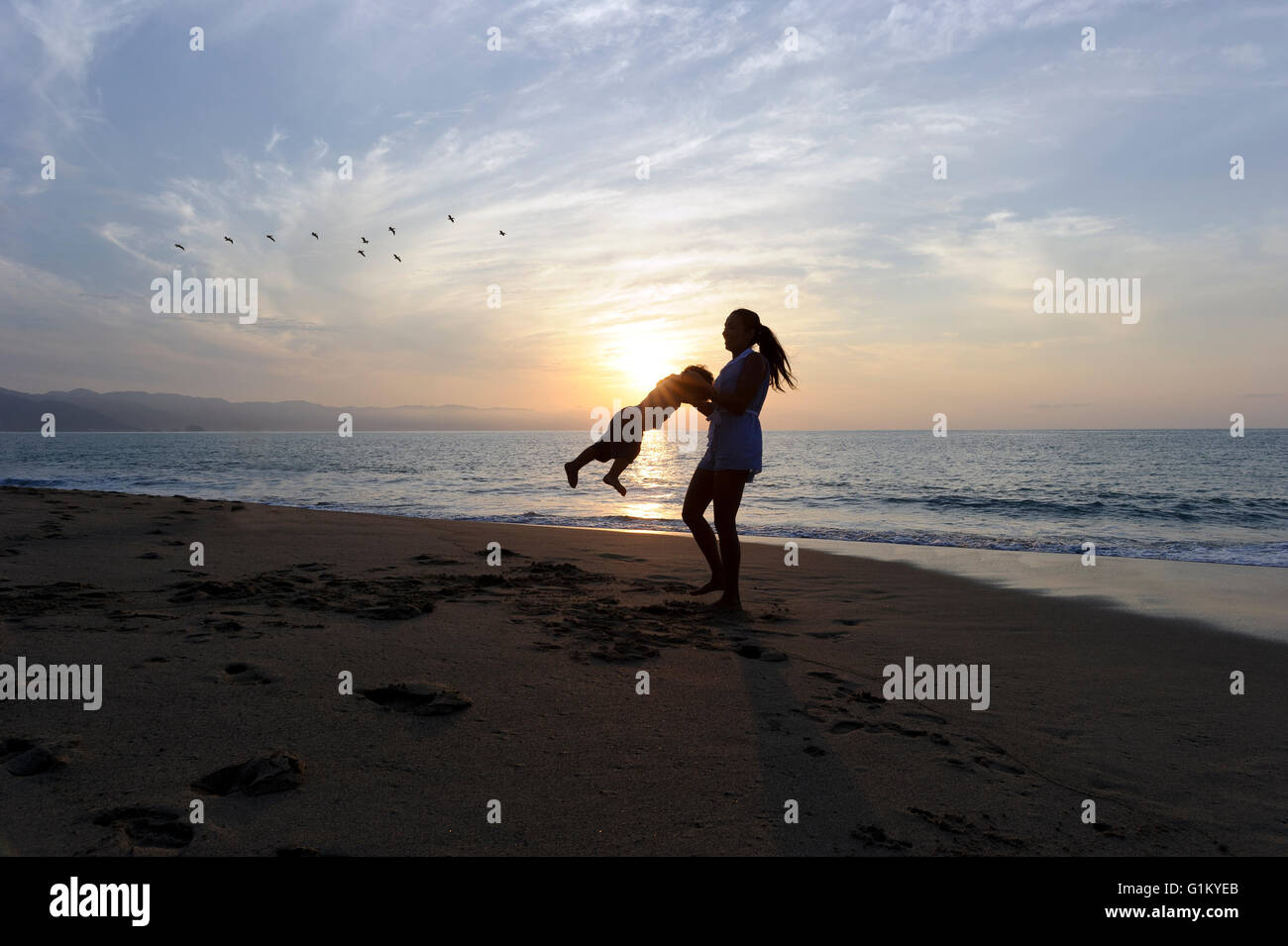 Mother son is a mother happily playing with her son as she swings him around on the beach as birds fly by in a sunset sky. Stock Photo