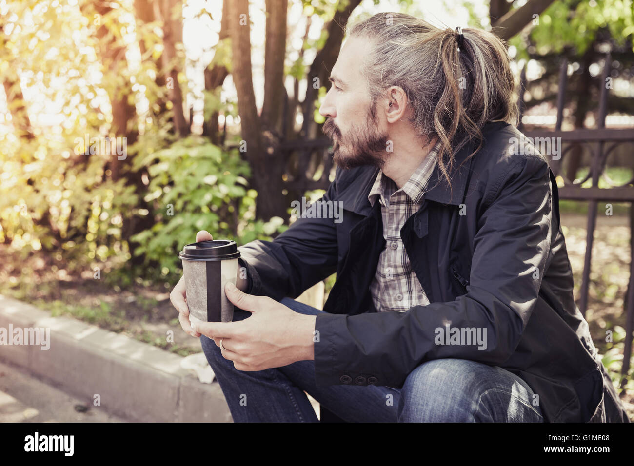 Bearded Asian man sitting on the sidewalk in the park with coffee in paper cup, outdoor portrait with selective focus and tonal Stock Photo
