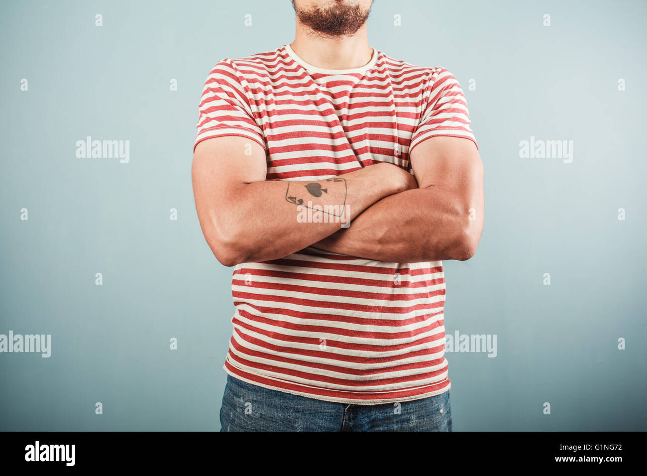 A young man with a tattoo is wearing a striped shirt and standing with his arms crossed Stock Photo