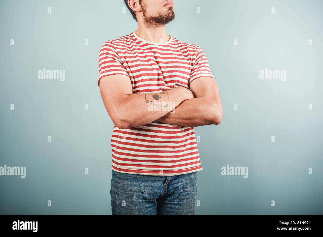 A young man with a tattoo is wearing a striped shirt and standing with his arms crossed Stock Photo
