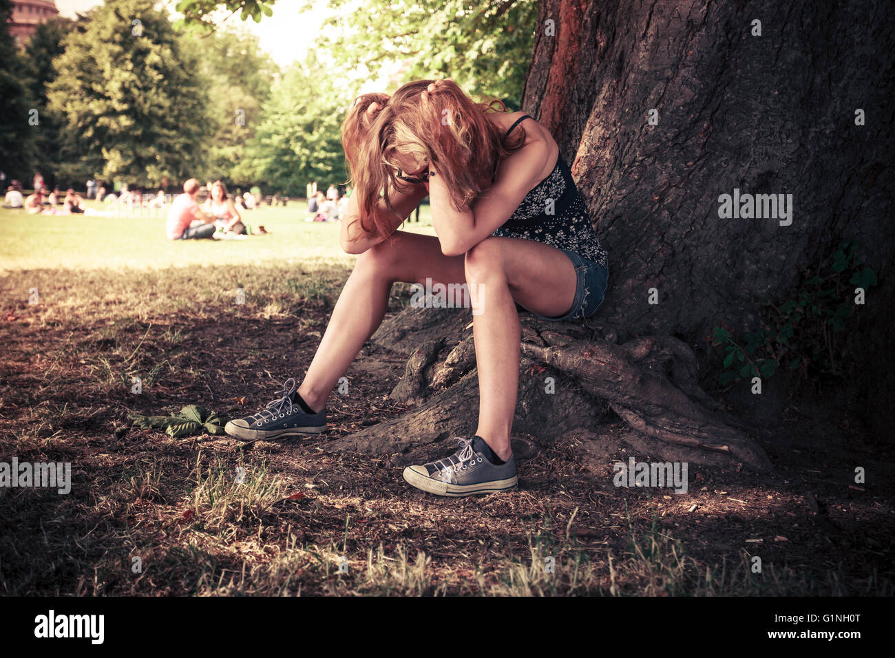 A tired an depressed young woman is sitting under a big tree in the park Stock Photo