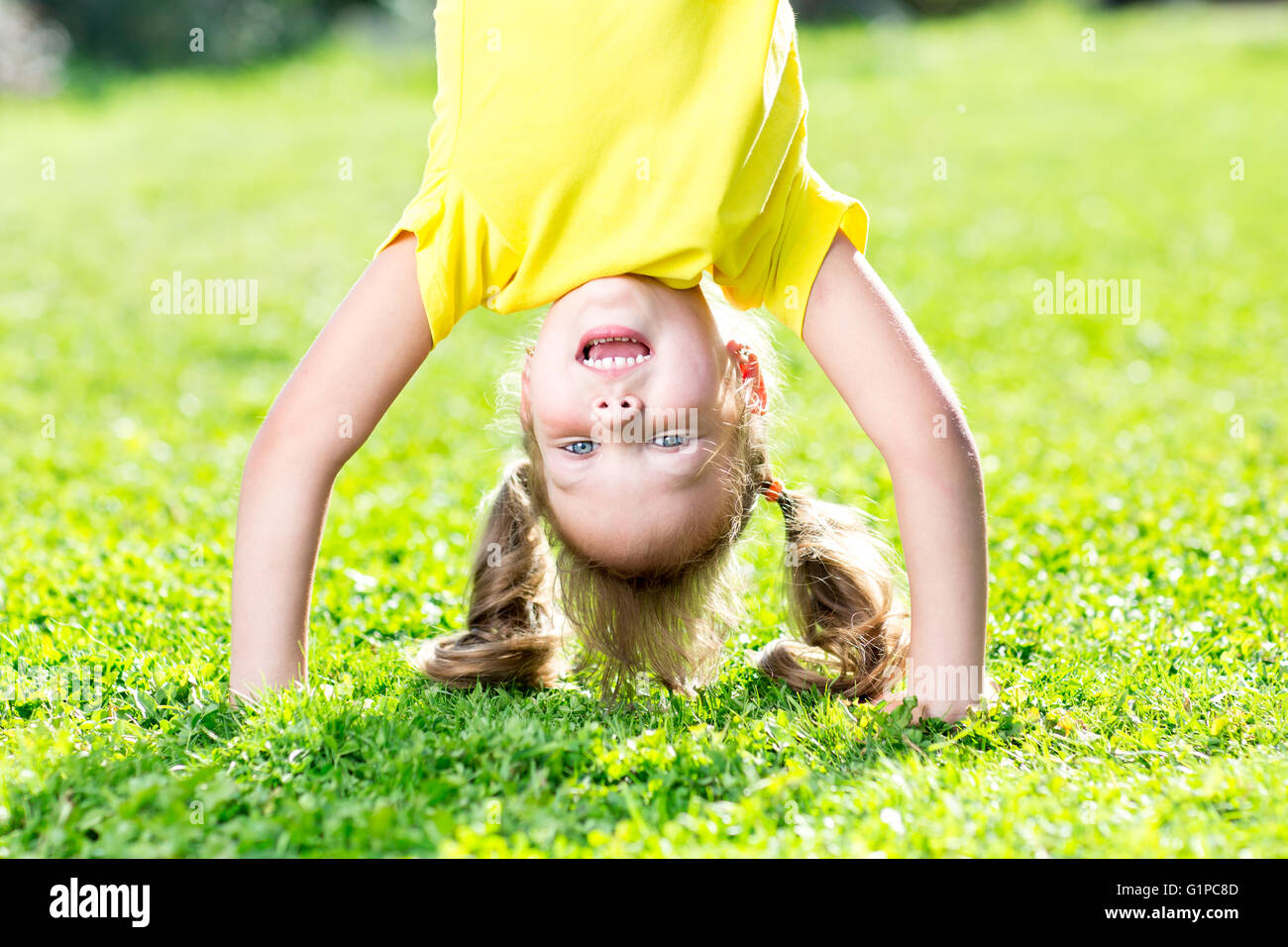 Happy little child girl have a fun standing on her head among grass ...