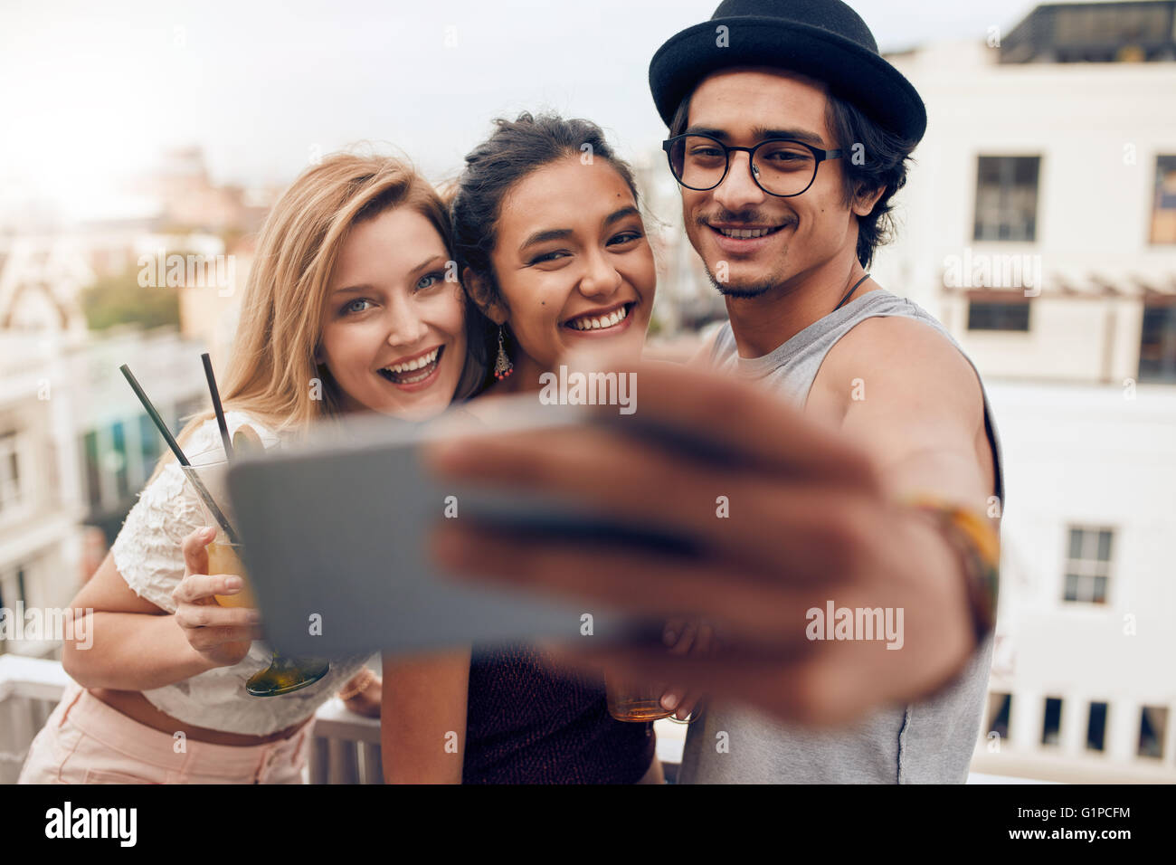 Shot of young man with female friends taking selfie on mobile phone. Young people partying on rooftop having fun. Stock Photo