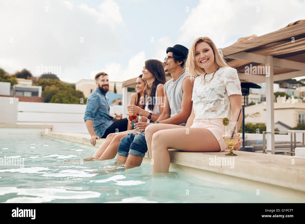 Portrait of happy young woman sitting by the pool with her friends partying. Young people relaxing by the swimming pool with the Stock Photo