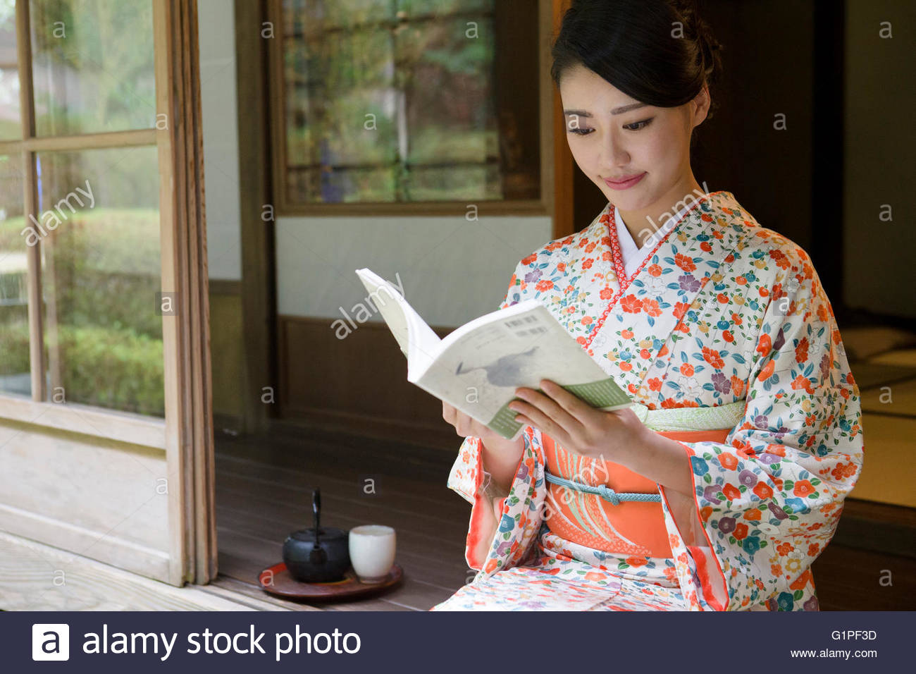 Young Japanese woman in kimono reading book sitting on wooden porch.