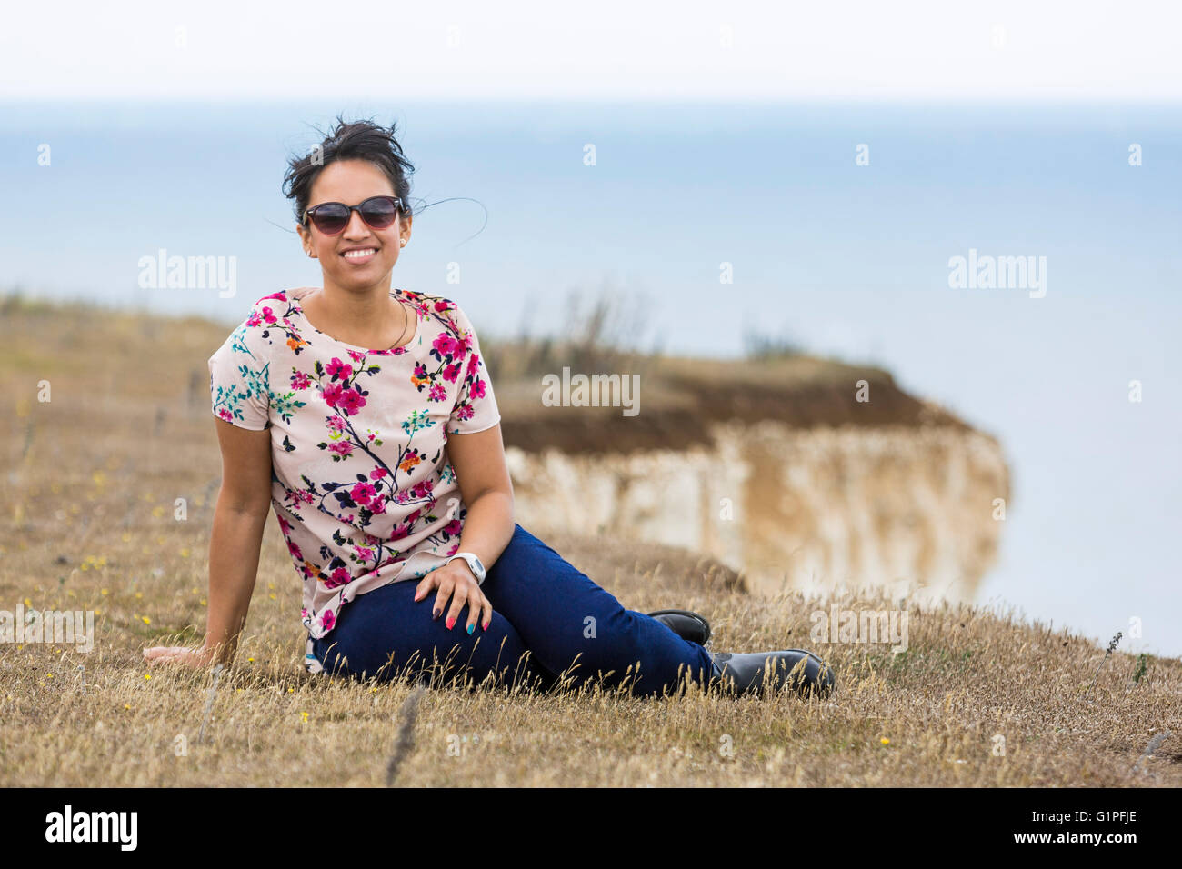 Young woman wearing sunglasses and sitting on the grass. The Seven Sisters Cliffs, South Down National Park, East Sussex, UK Stock Photo