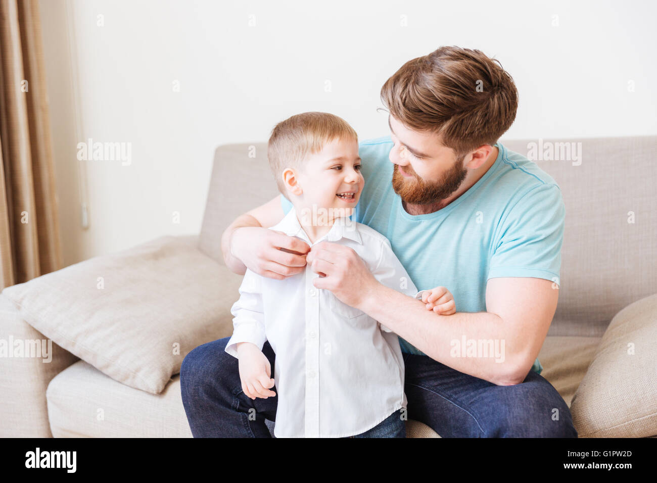 Happy father sitting on sofa and dressing his little son at home Stock Photo