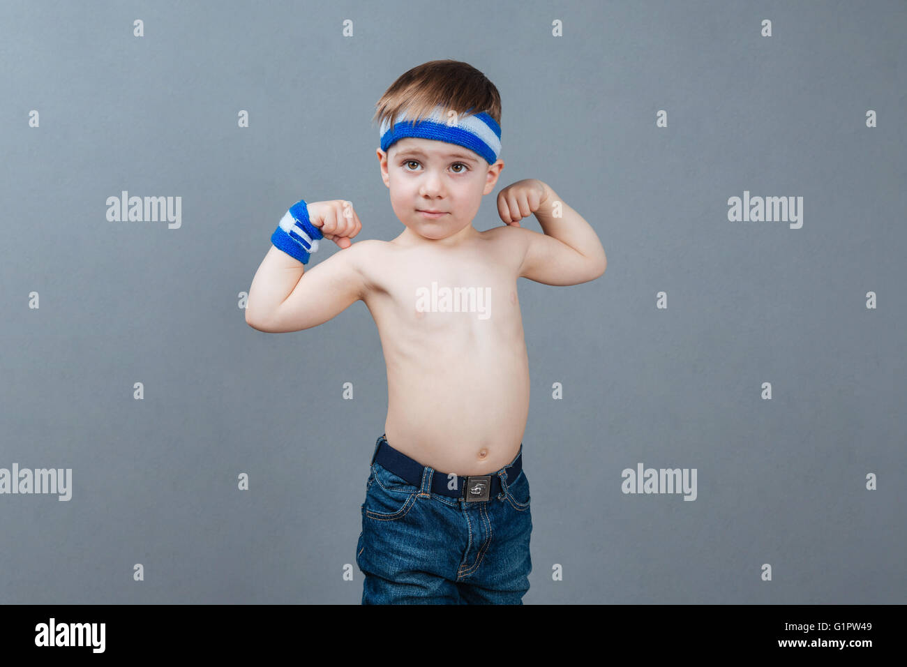 Portrait of shirtless little boy standing and showing biceps over grey