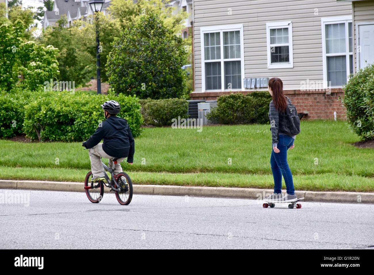 Two teens riding a bike and skateboard down a residential street Stock Photo