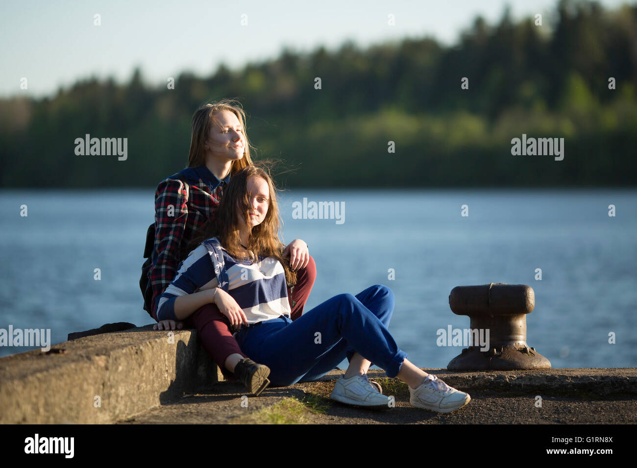 Two friends teen girls spend time together at the pier of the river. Stock Photo