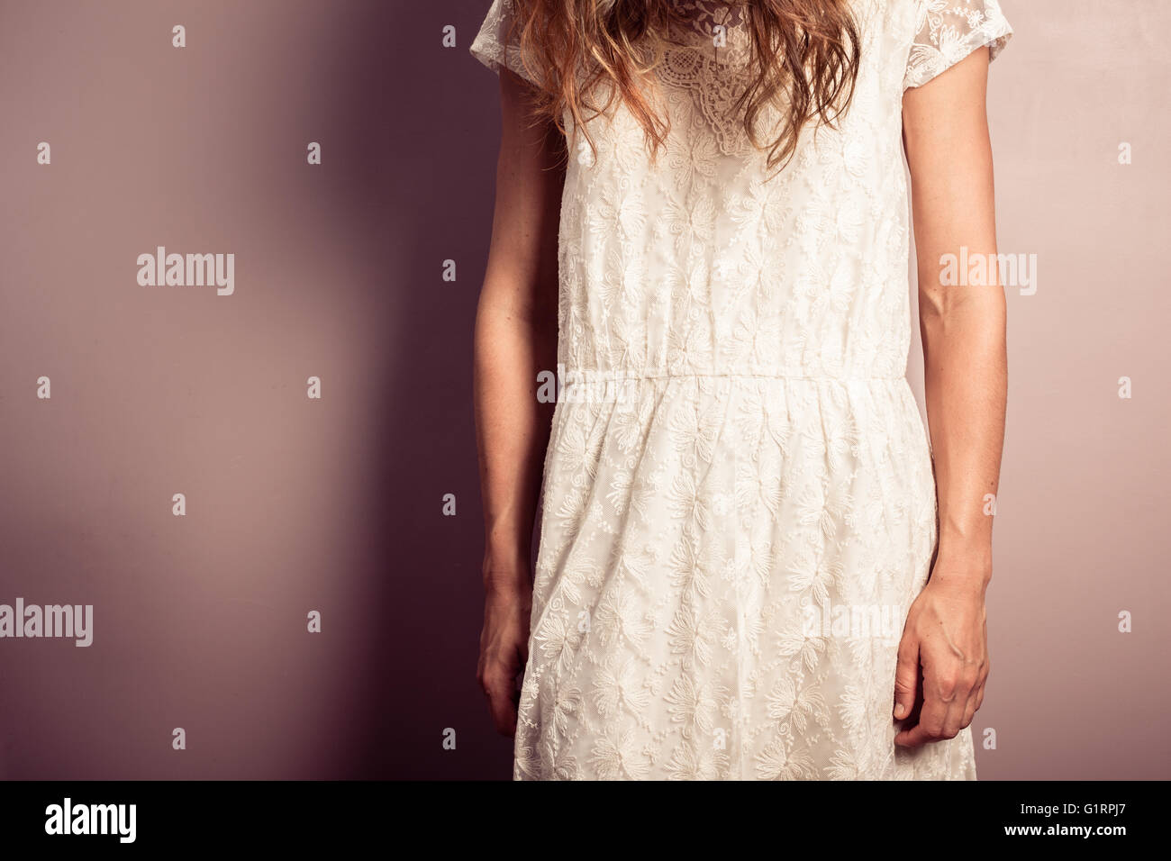 A sad young woman wearing a white dress is standing by a purple wall with her head hanging Stock Photo