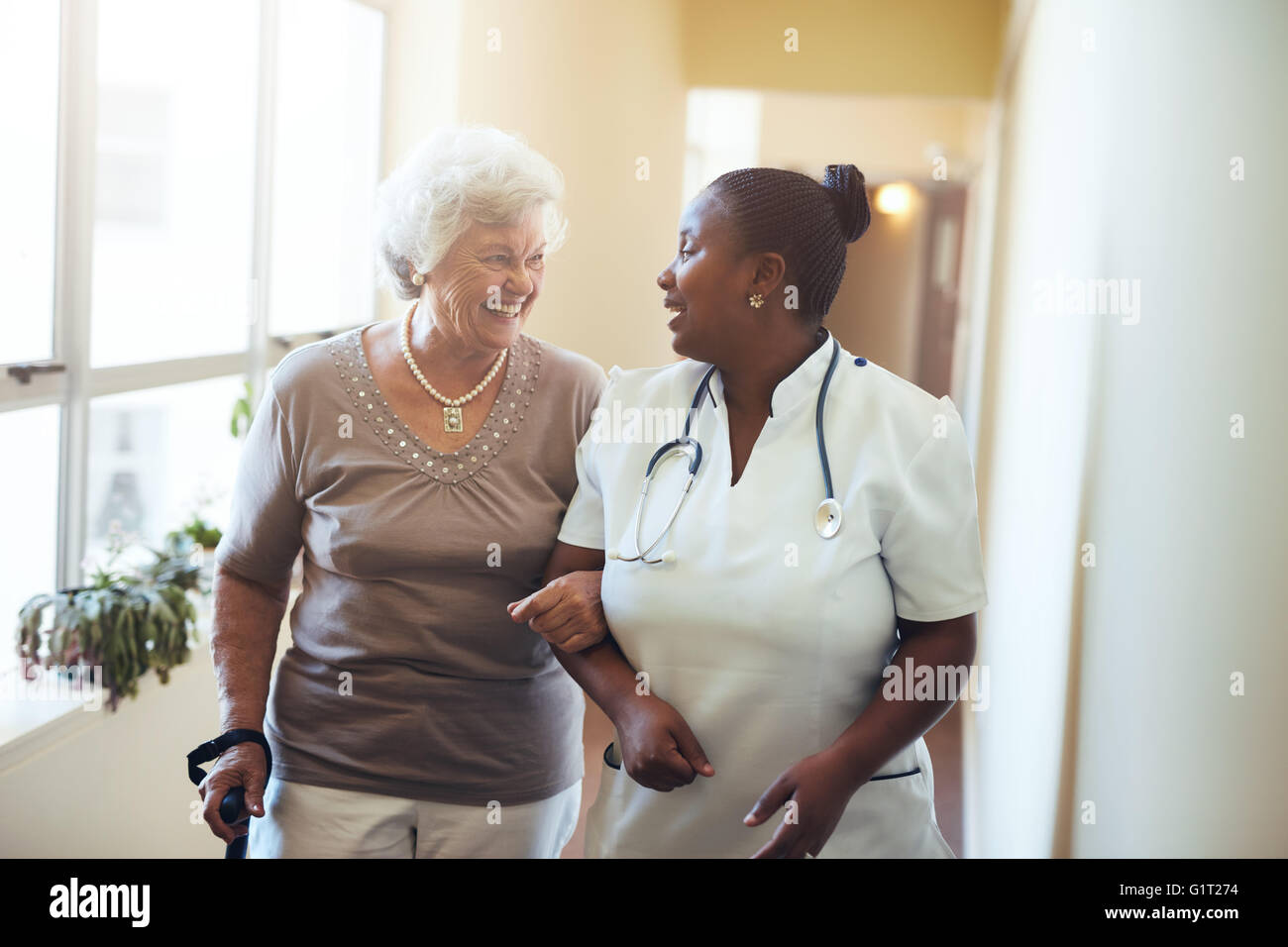 Senior woman walking in the nursing home supported by a caregiver. Nurse assisting senior woman. Stock Photo