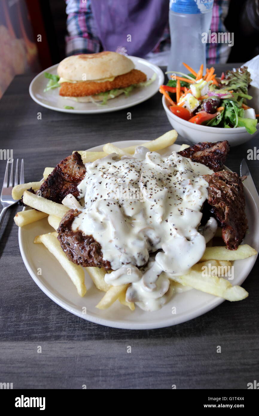 A plate of  steak with mushroom sauce and chips with a bowl of Greek salad and Chicken Schnitzel burger in the background Stock Photo