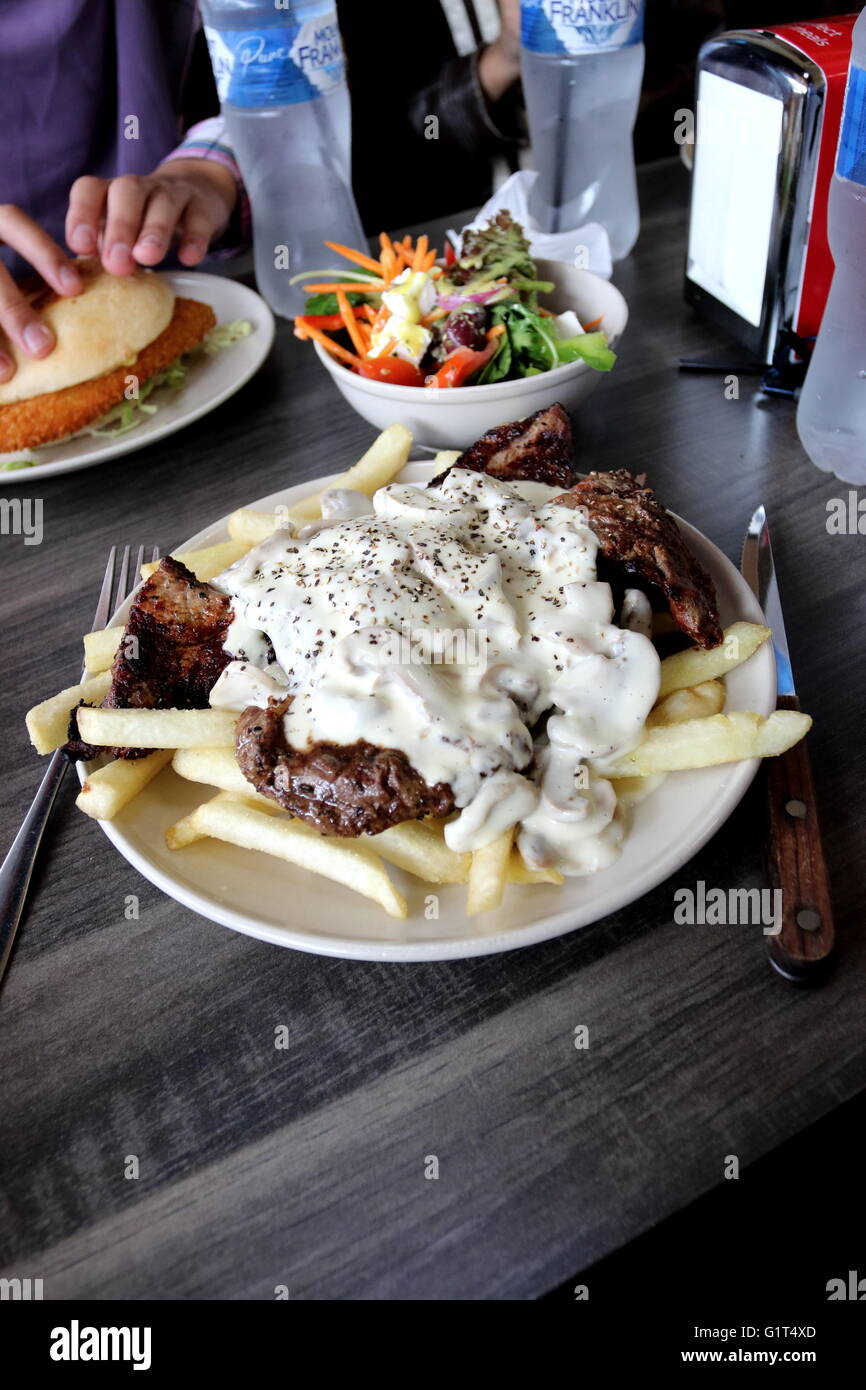 A plate of  steak with mushroom sauce and chips with a bowl of Greek salad and Chicken Schnitzel burger in the background Stock Photo