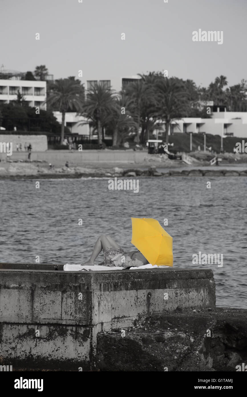 Someone sun bathing under a bright yellow umbrella on a concrete harbour Stock Photo