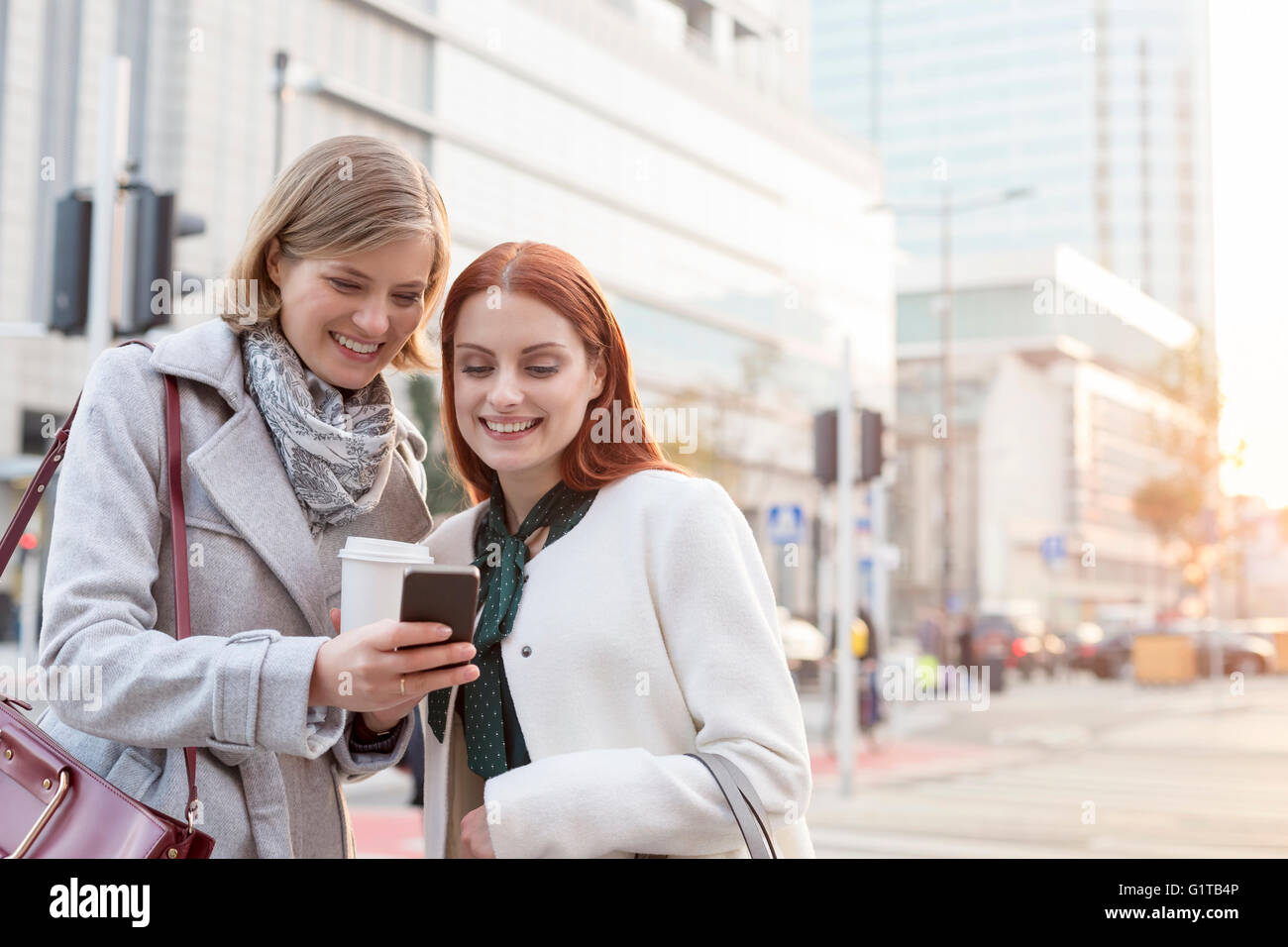 Smiling businesswomen texting and drinking coffee on city street Stock Photo
