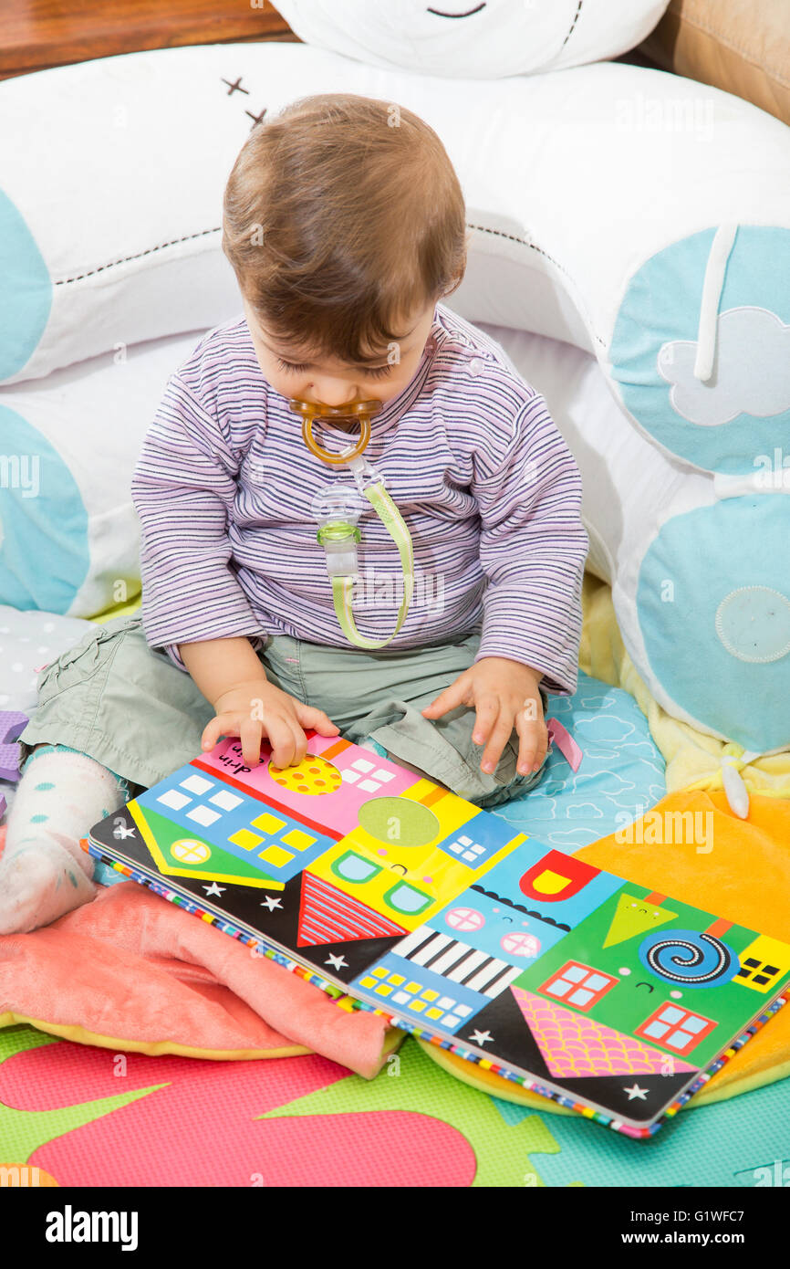 Little one year old baby sitting on floor and looking at bright colorful book with pictures Stock Photo