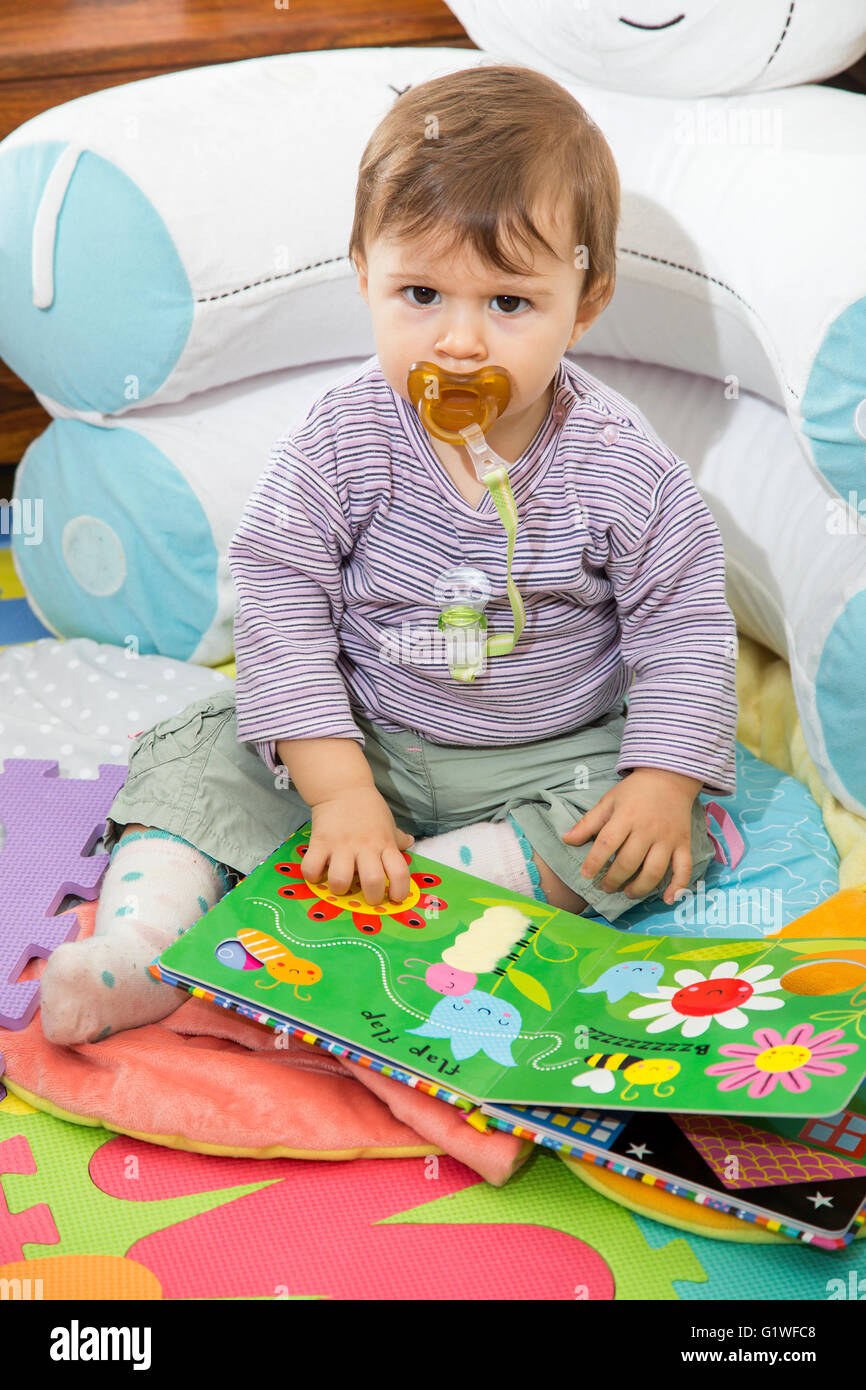 Little one year old baby with colorful book sitting on floor against of pillows and looking seriously at camera Stock Photo