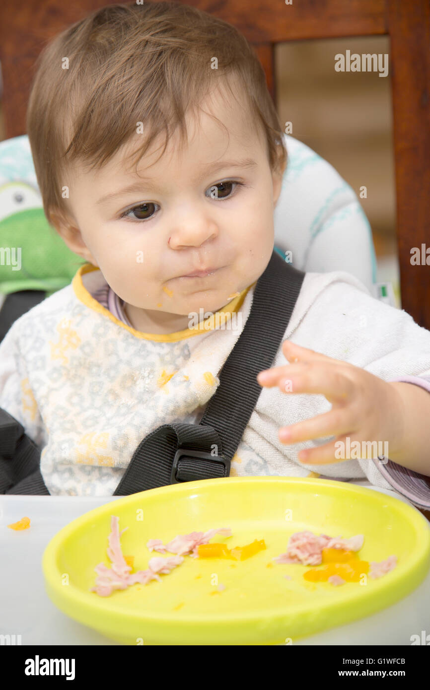 Portrait of cute one year old baby with dirty shirt eating with hands Stock Photo