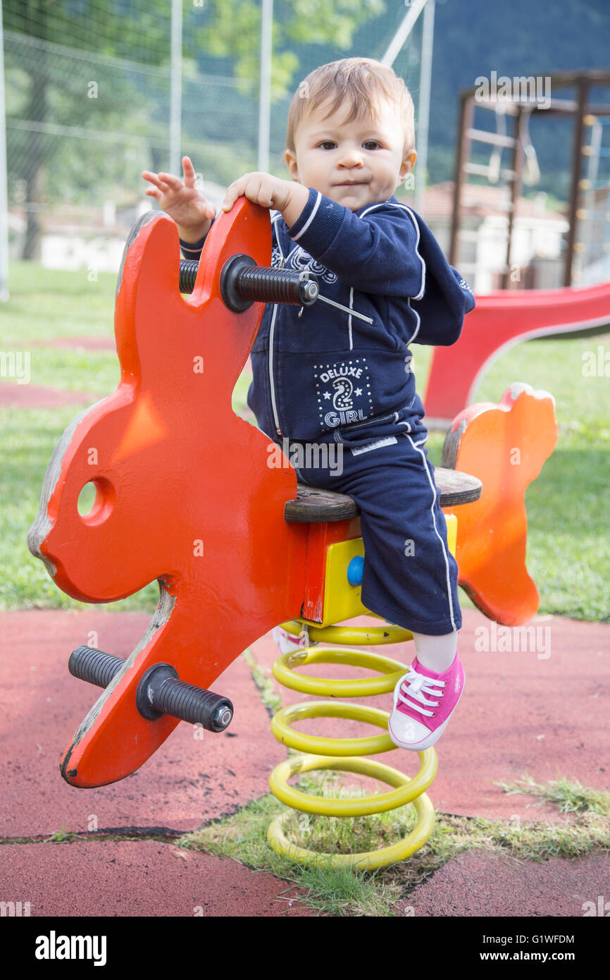 Portrait of lovely serious one year old baby sitting on colorful swing and looking at camera at playground Stock Photo