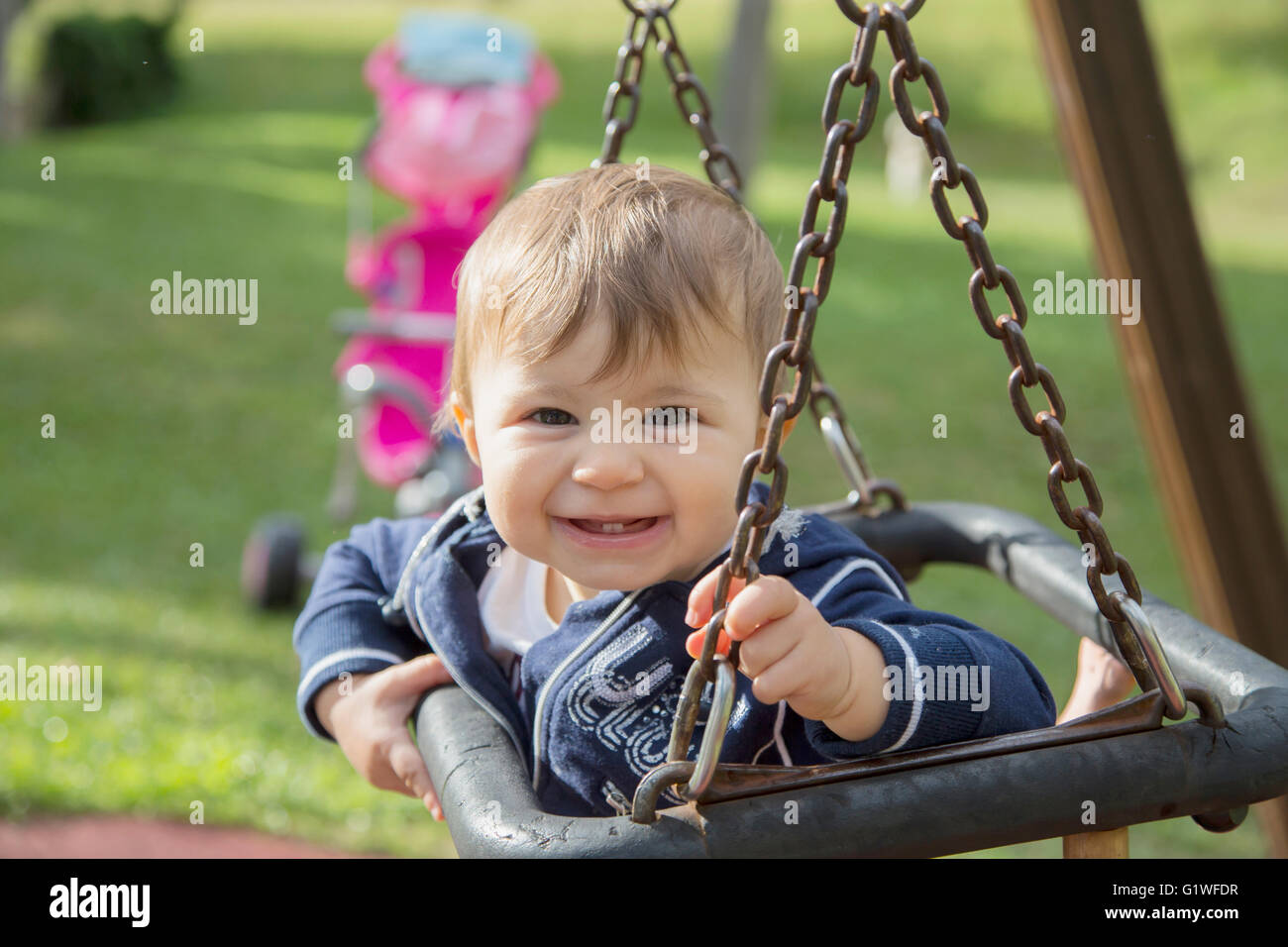 Lovely one year old kid smiling while sitting on swing at playground Stock Photo
