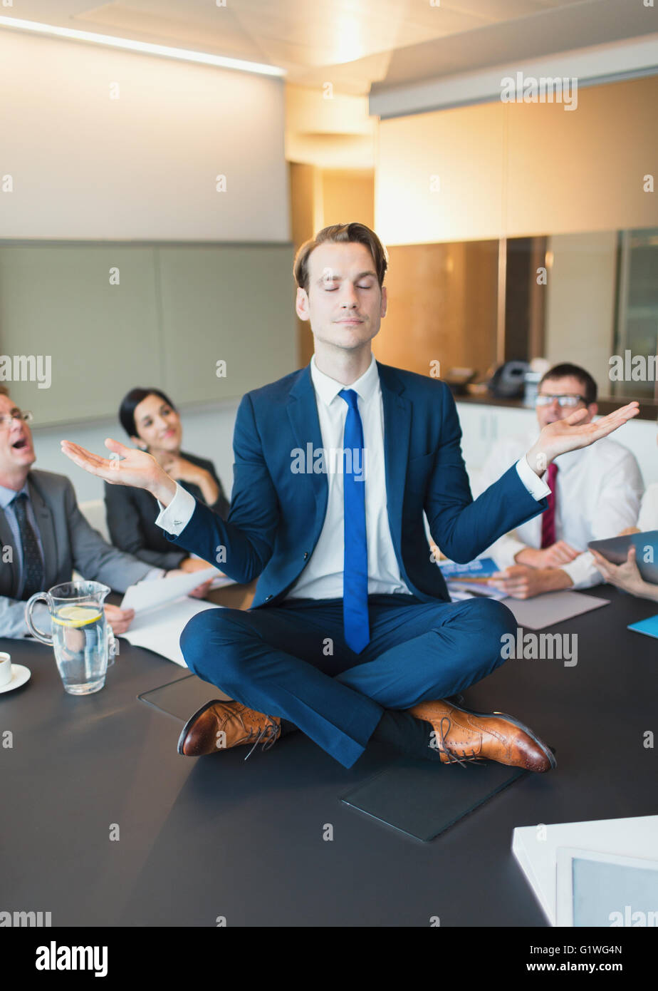 Colleagues watching zen-like businessman meditating in lotus position on conference table Stock Photo