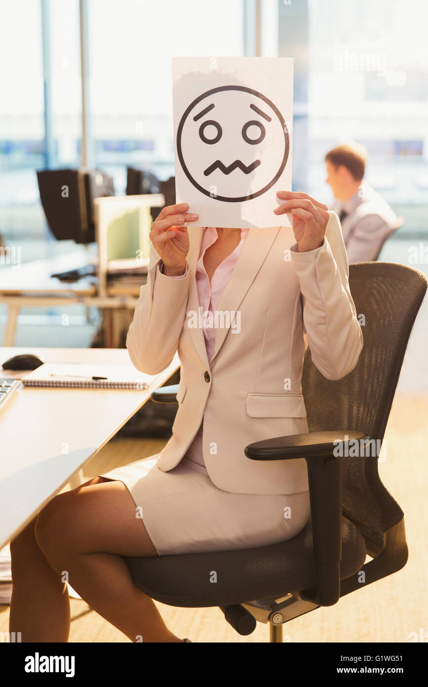 Portrait of businesswoman holding frowning face printout over her face in office Stock Photo