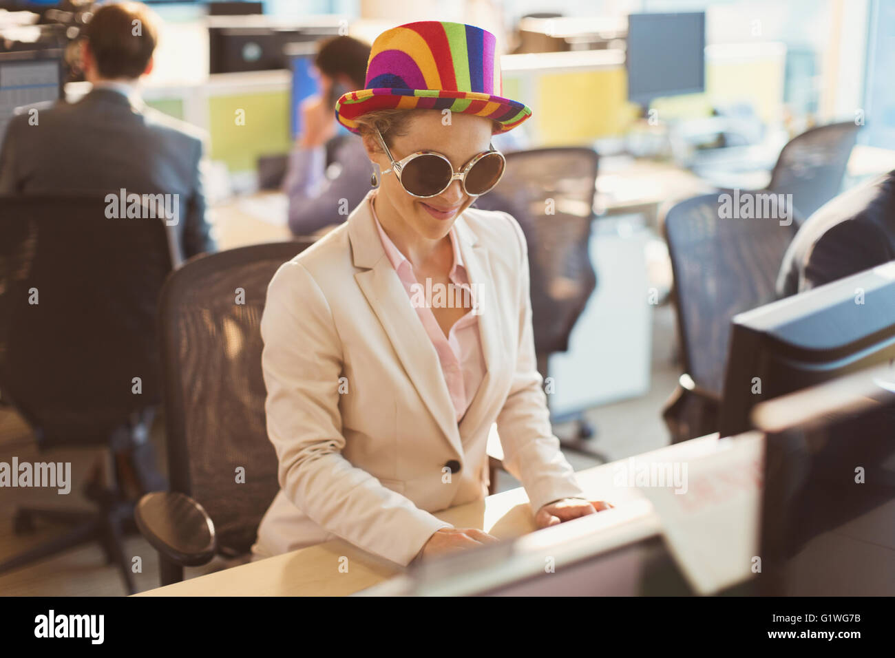Playful businesswoman wearing silly sunglasses and striped hat at computer in office Stock Photo