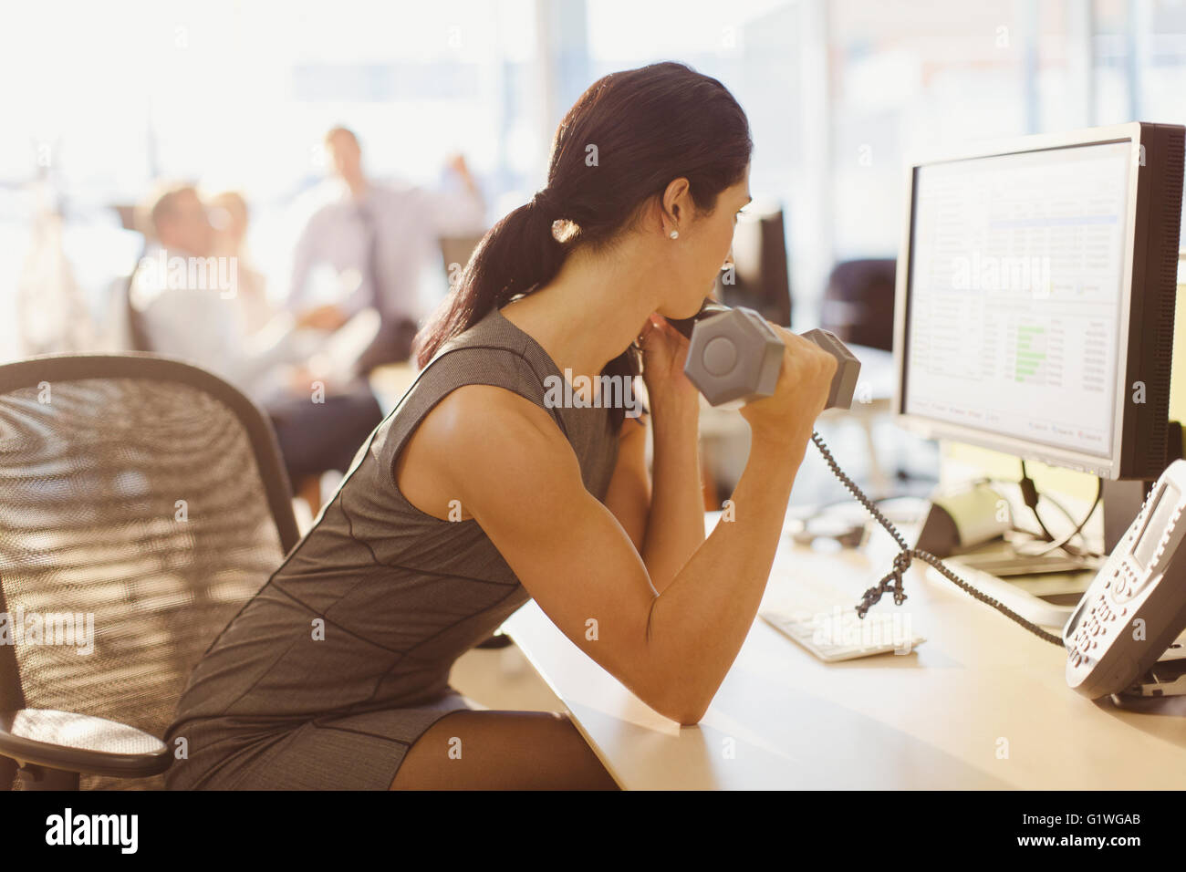 Businesswoman doing biceps curls with dumbbell and talking on telephone at computer in office Stock Photo