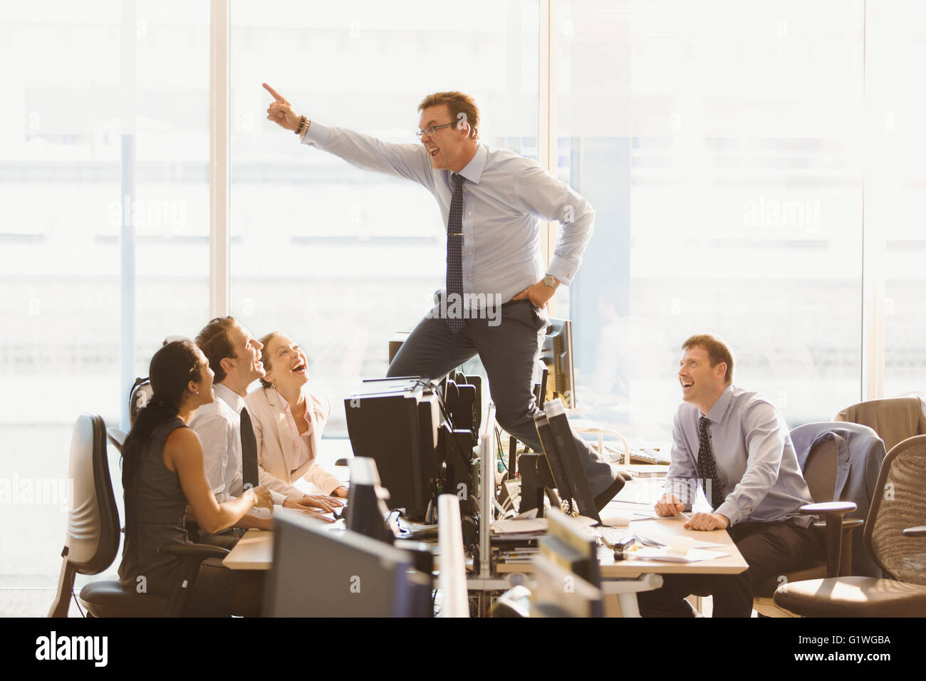 Laughing colleagues watching exuberant businessman dancing on top of desks in office Stock Photo