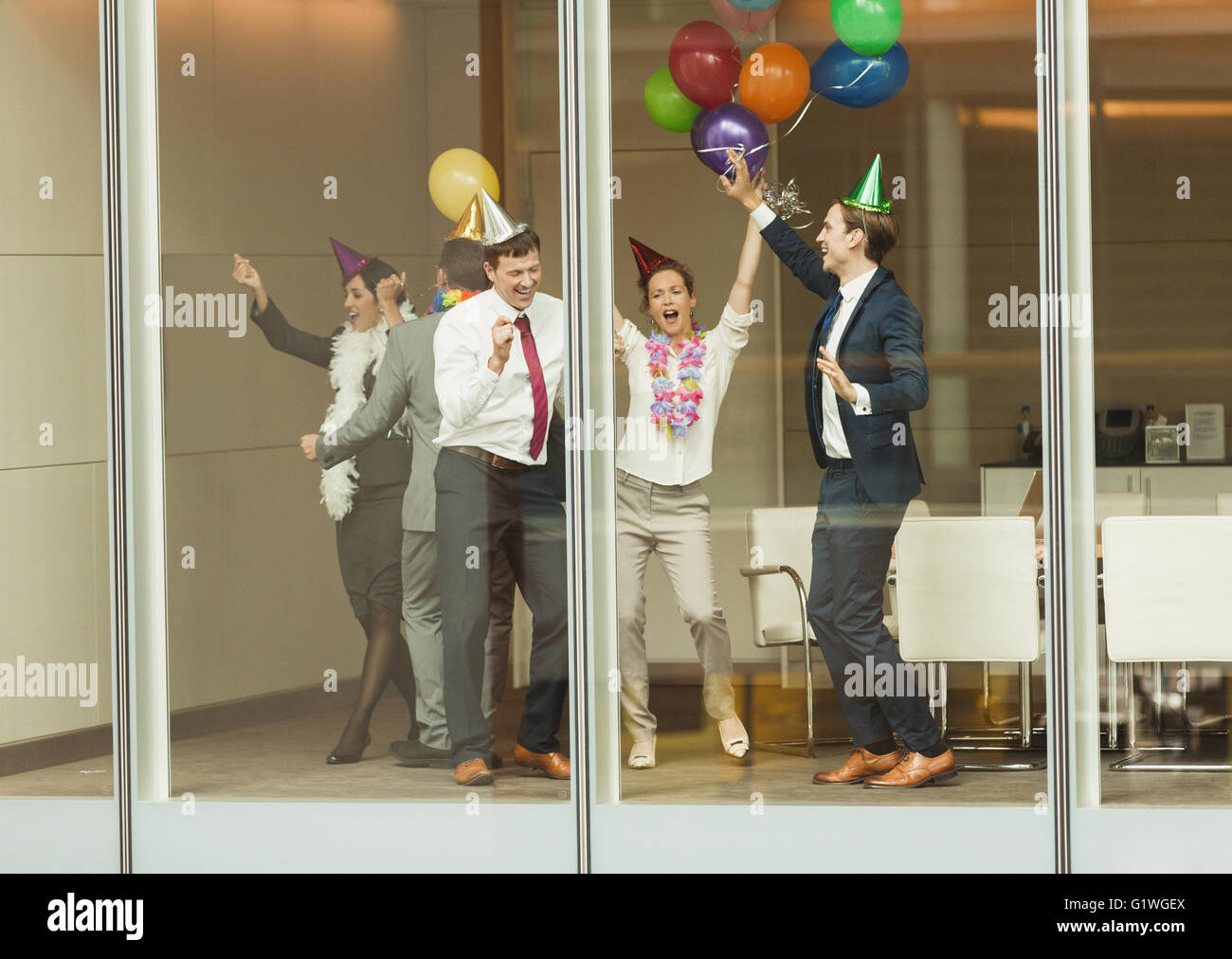 Business people wearing party hats and dancing with balloons at conference room window Stock Photo