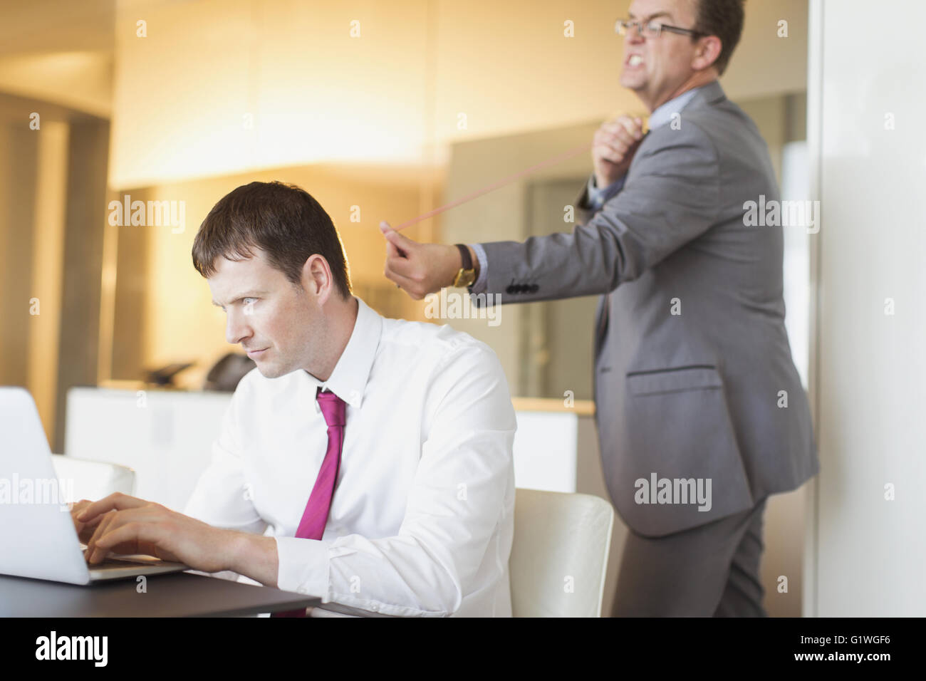 Angry businessman aiming rubber band at unsuspecting businessman working at laptop Stock Photo