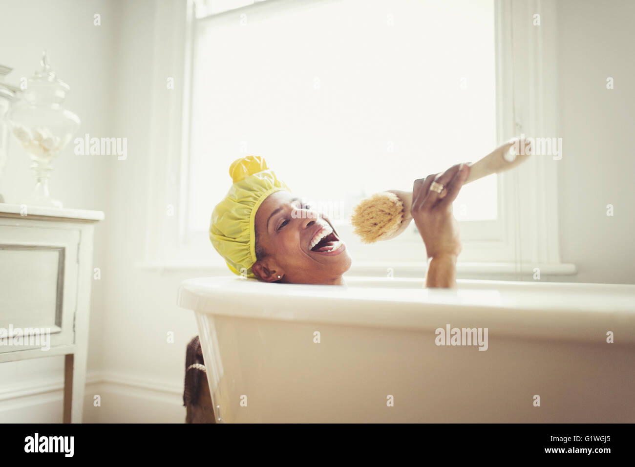 Playful mature woman singing into loofah brush in bathtub Stock Photo