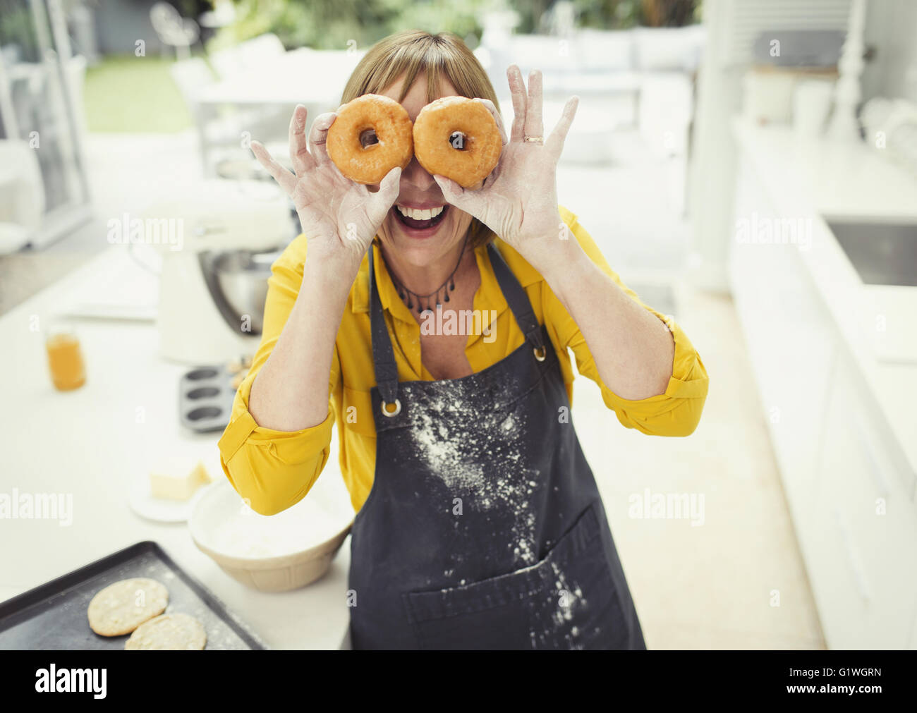 Portrait playful mature woman covering eyes with donuts in kitchen Stock Photo