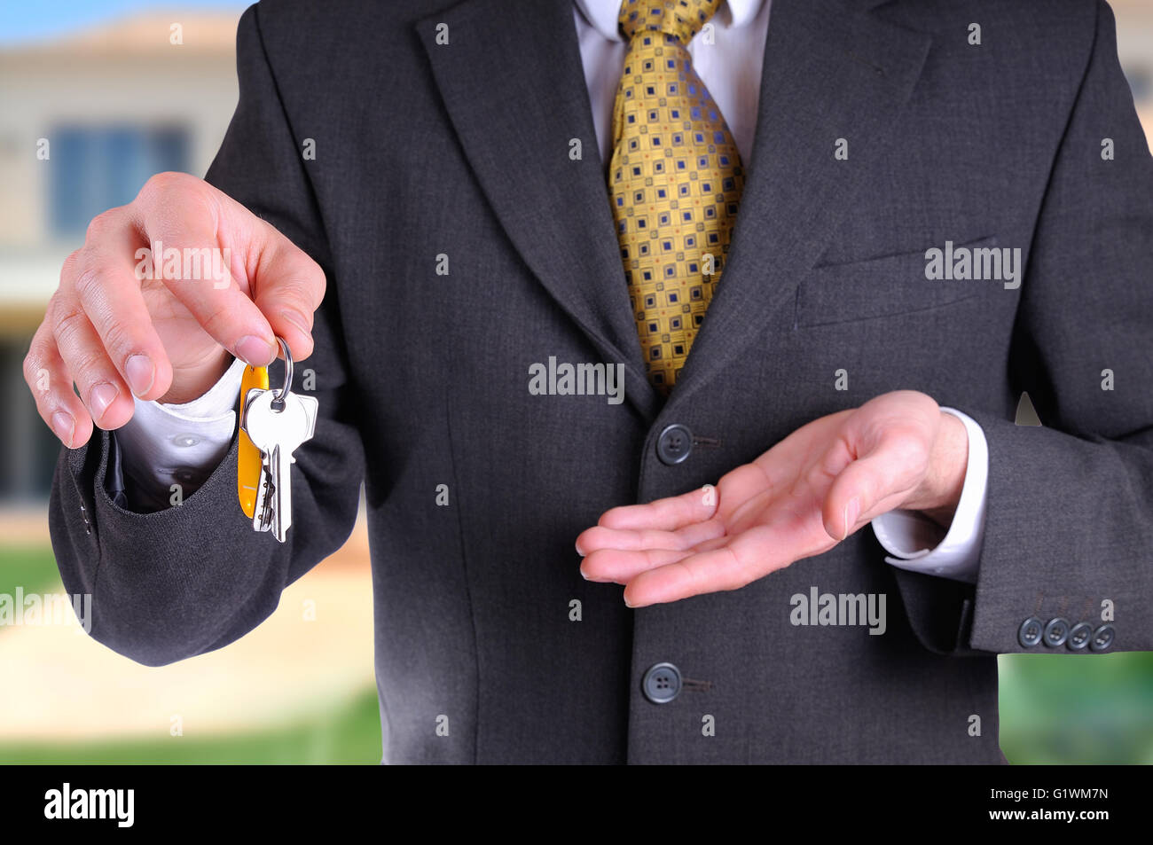 Front half-body commercial agent wearing a dark gray costume handing the keys for the purchase of a house with home background Stock Photo