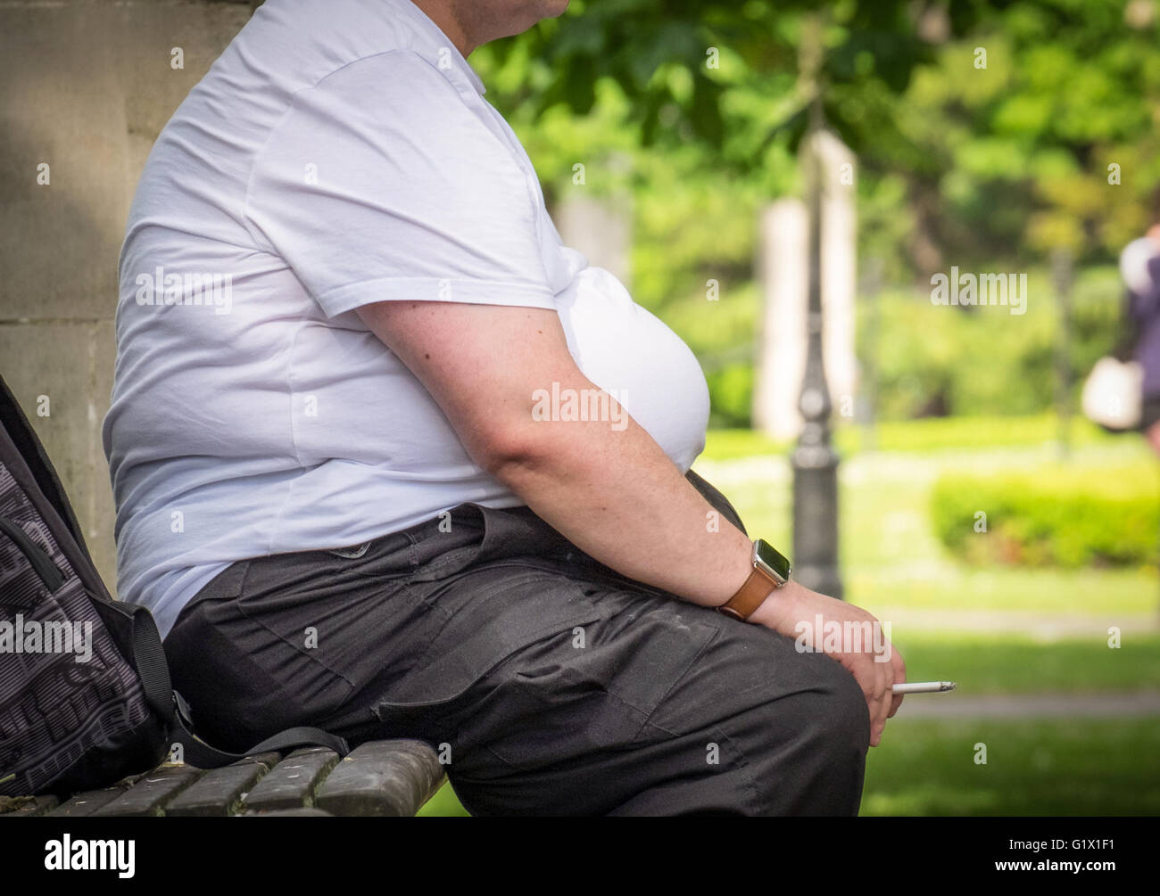 Photo of an overweight obese man smoking whilst sitting on a park bench in the UK Stock Photo