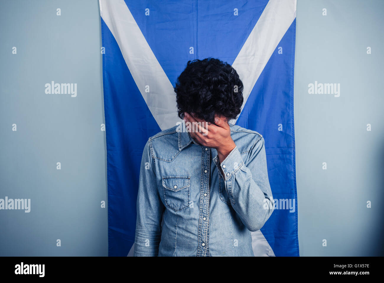 A depressed man is standing in front of a Scottish flag with his head in his hands Stock Photo