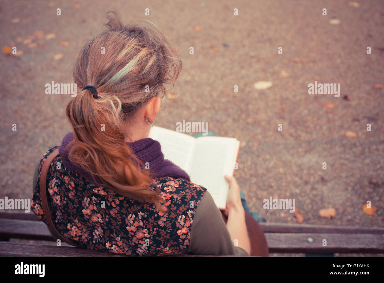 A young woman is sitting on a park bench in the autumn and is reading a ...