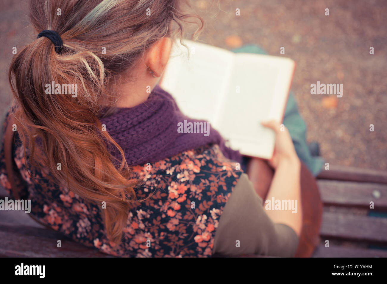 A young woman is sitting on a park bench in the autumn and is reading a ...