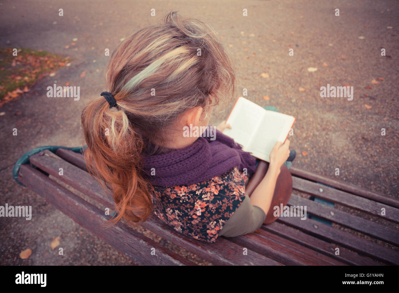 A young woman is sitting on a park bench in the autumn and is reading a ...