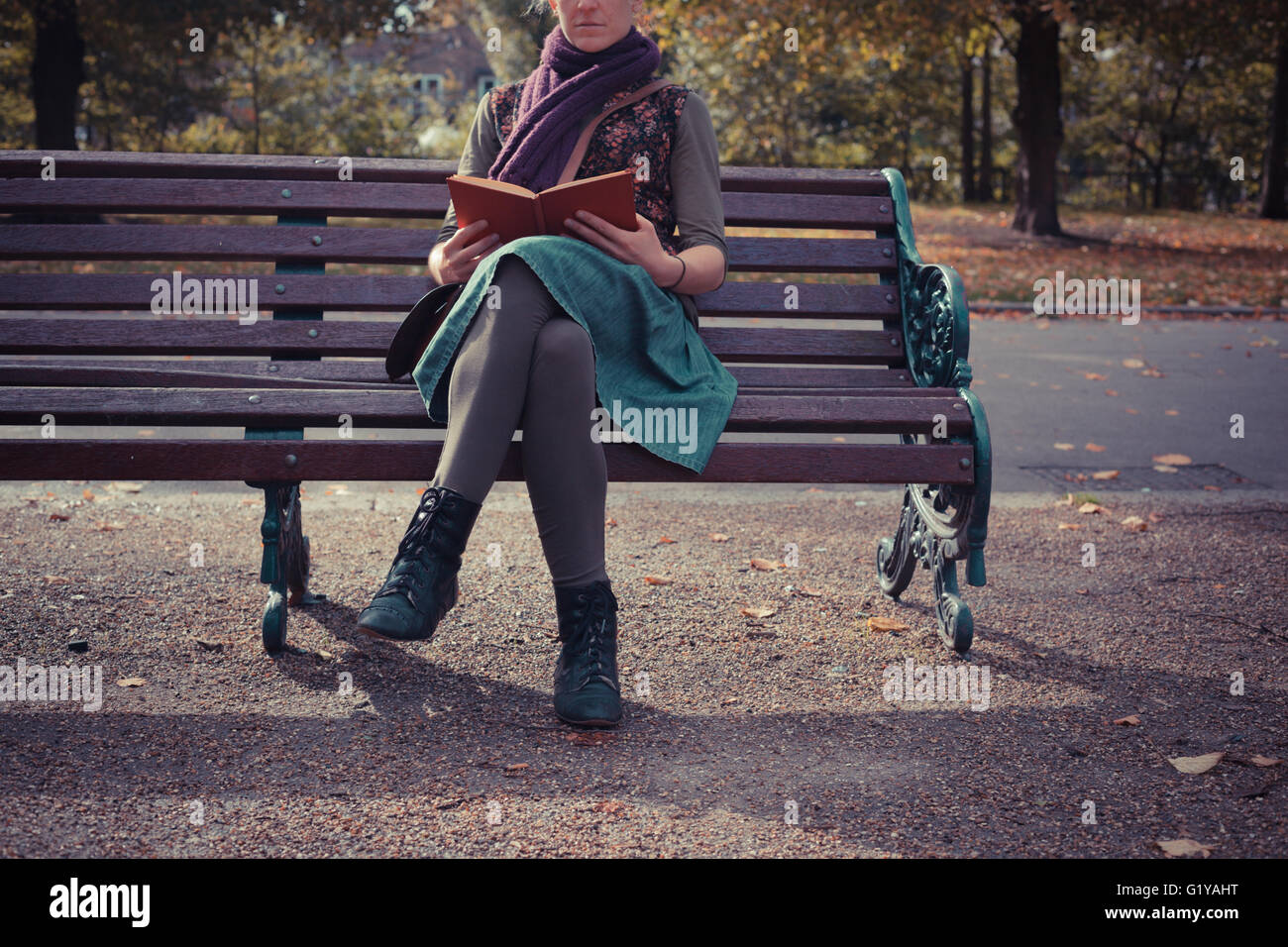 A young woman is sitting on a park bench in the autumn and is reading a ...
