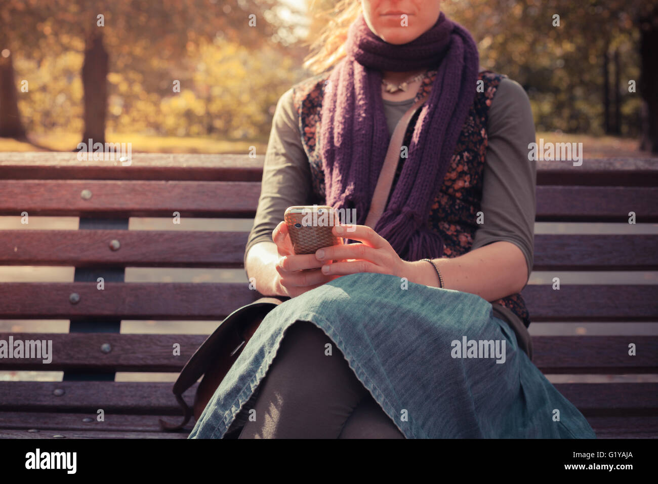 A young woman is sitting on a park bench in autumn and is using her ...