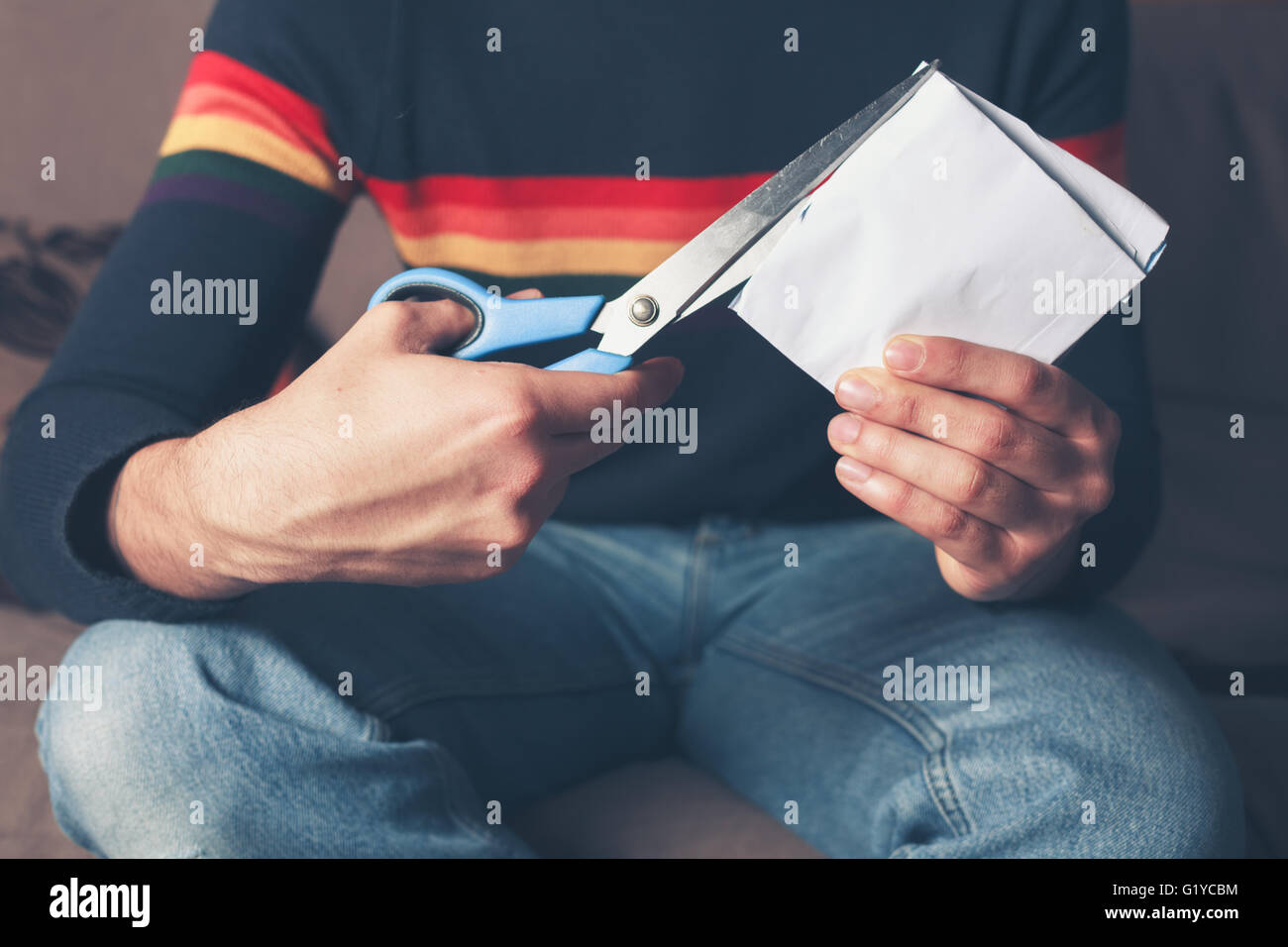A young man is sitting on a sofa and is cutting a piece of paper with