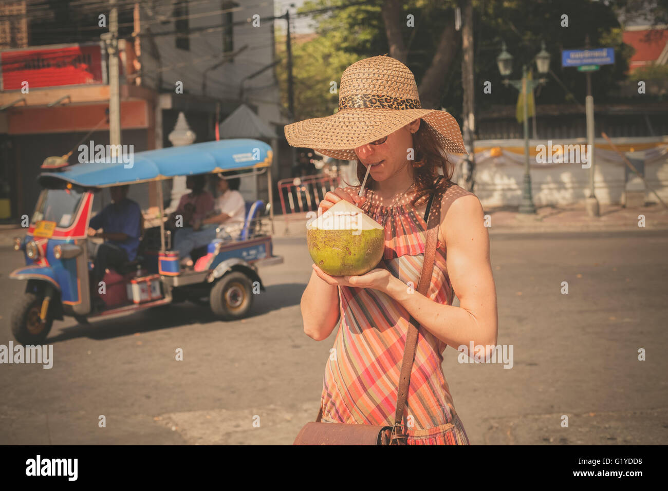 A young woman is drinking from a coconut in the street of an Asian country with a tuktuk in the background Stock Photo
