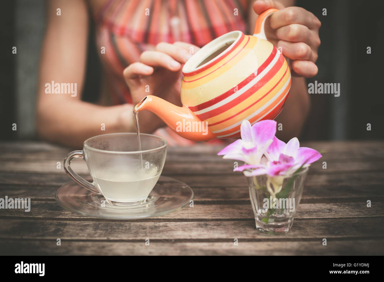 A young woman is sitting at a table and is pouring a cup of tea Stock Photo