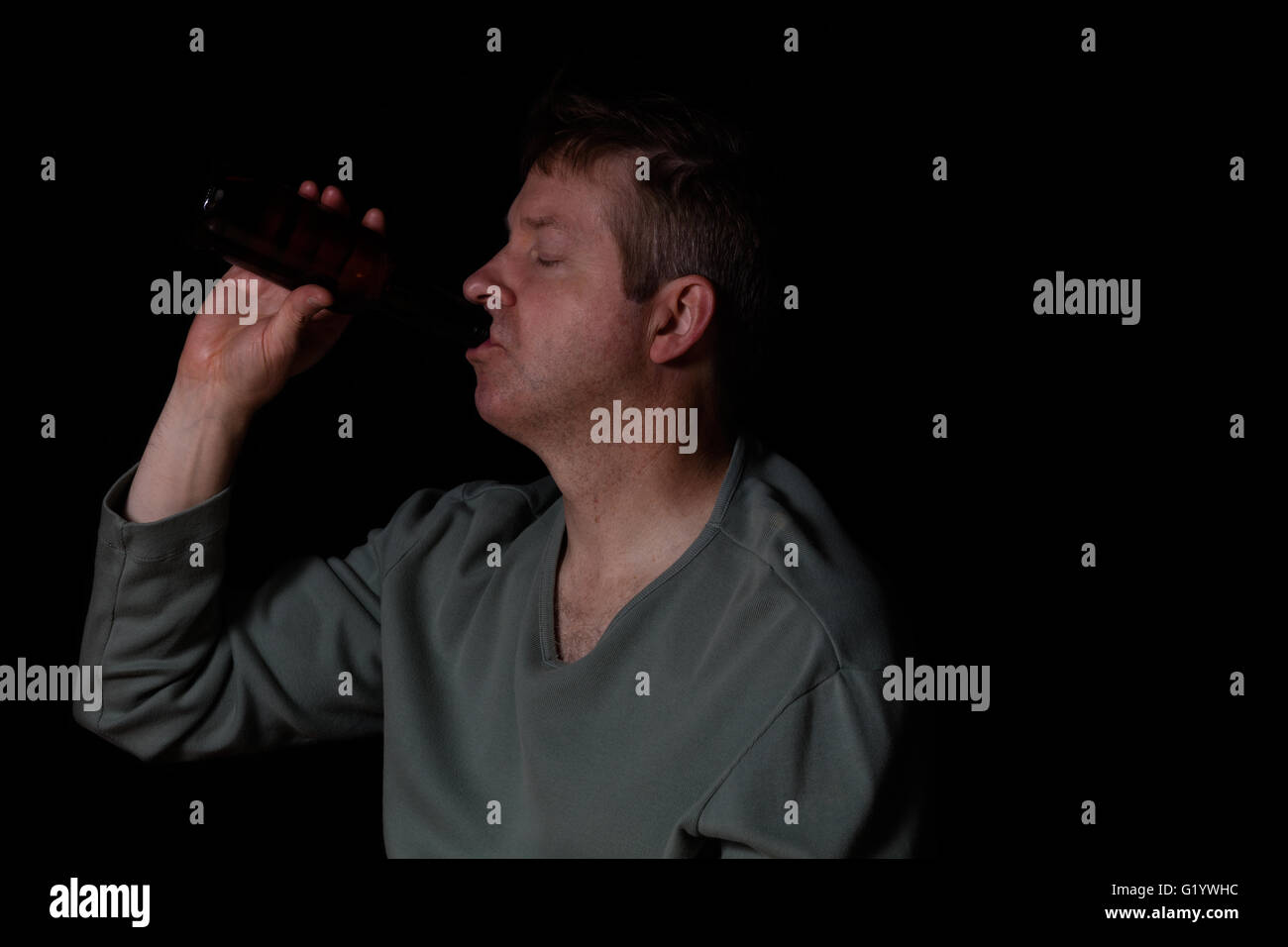 Depressed man drinking beer out of a bottle while sitting down with eyes close in a dark background. Stock Photo