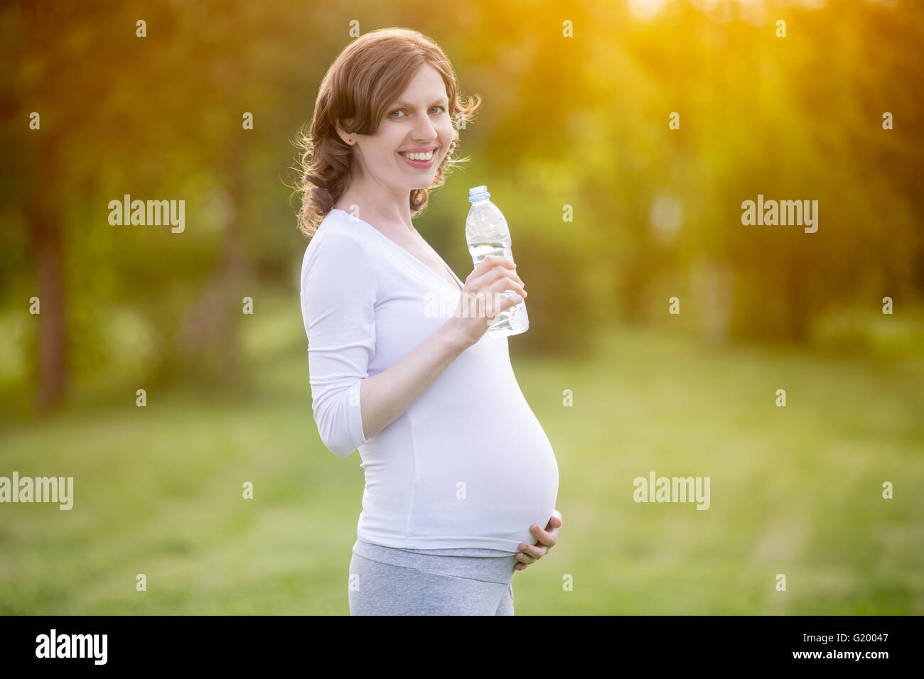 Portrait of happy young pregnant model standing with bottle of fresh water and looking at camera. Future mom expecting baby Stock Photo