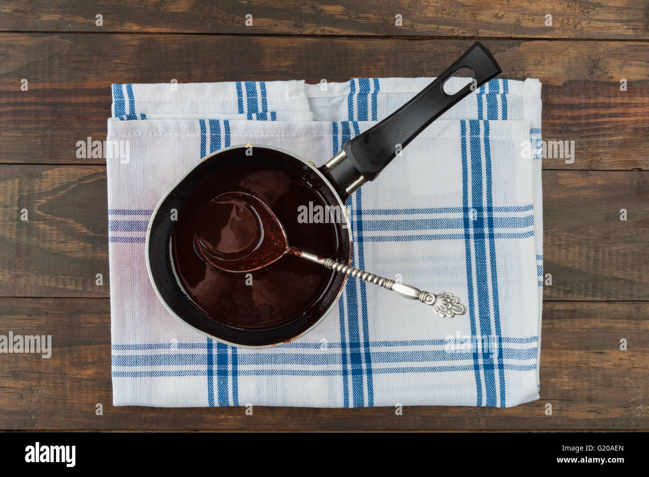 Pan filled with dark melted chocolate with vintage silver spoon on checkered napkin and wooden table. Top view Stock Photo