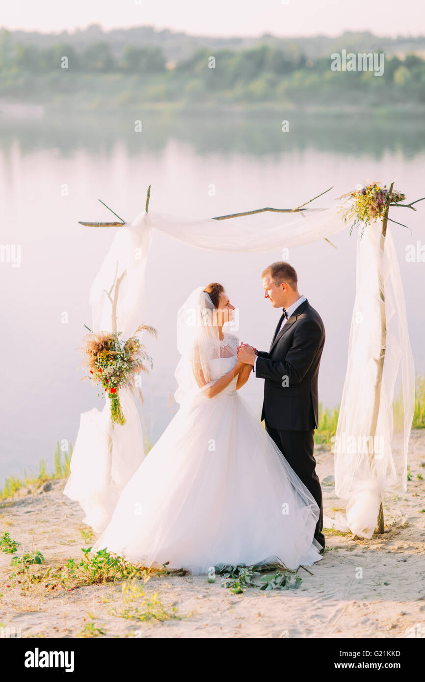Beautiful bride in white dress and handsome groom wearing black suit standing holding hands under archway on beach wedding ceremony near lake Stock Photo - Alamy Beautiful bride in white dress and handsome groom wearing black suit standing holding hands under archway on beach wedding ceremony near lake Stock Photo - Alamy